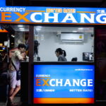 Man in gray short standing in front a counter of currency exchange.